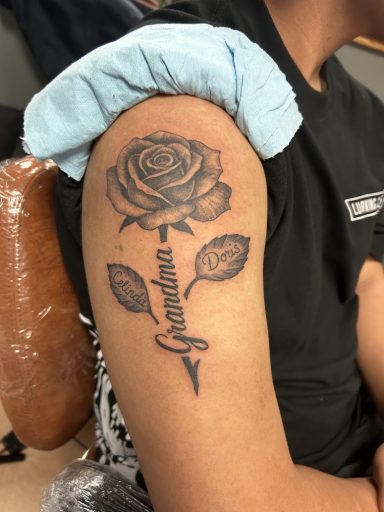 Black and grey realism tattoo of a rose on a shoulder with the name Grandma and a date in elegant script.